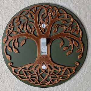 May include: A decorative light switch cover featuring a bronze-colored tree of life design on a green background. The tree's branches and roots are intricately detailed. The switch is white, with "ON" printed on it.