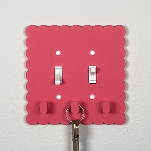 May include: A square, hot pink light switch cover plate with a scalloped edge. The plate features two white toggle light switches, four white mounting screws, and three small, rounded hooks. A silver metal key ring hangs from the center hook.