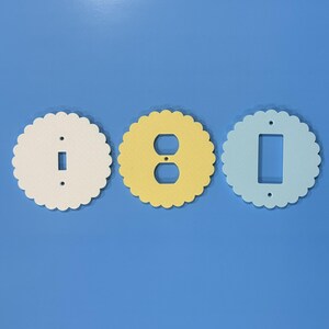 May include: Three decorative light switch covers in a scalloped circle design. One is off-white with a single switch opening, another is yellow with a double outlet opening, and the third is light blue with a rectangular switch opening.