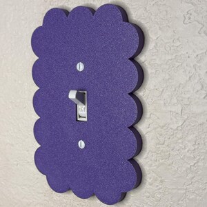 May include: A decorative purple light switch cover with a scalloped edge. The cover has a textured surface and a standard toggle switch. Two screws secure the cover to the wall. The switch is in the "on" position.