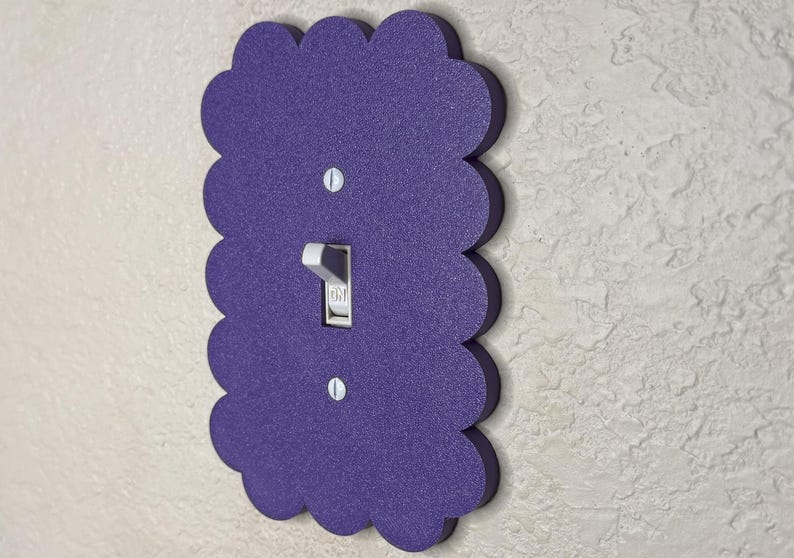 May include: A decorative purple light switch cover with a scalloped edge. The cover has a textured surface and a standard toggle switch. Two screws secure the cover to the wall. The switch is in the "on" position.