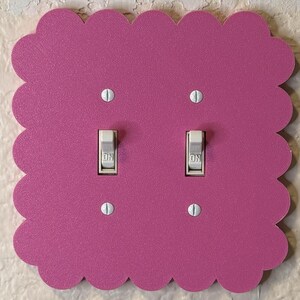 May include: A decorative, scalloped-edge light switch cover in a vibrant magenta. The cover features two light switches with the word "ON" visible. The cover is mounted on a textured, off-white wall.
