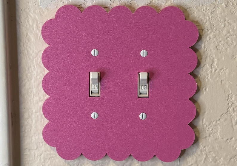 May include: A decorative, scalloped-edge light switch cover in a vibrant magenta. The cover features two light switches with the word "ON" visible. The cover is mounted on a textured, off-white wall.