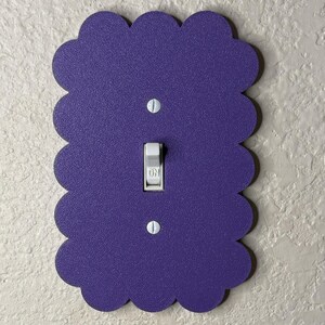 May include: A decorative purple light switch cover with a scalloped edge. The cover has a standard toggle switch in the center, with two screws at the top and bottom. The switch is in the "ON" position.