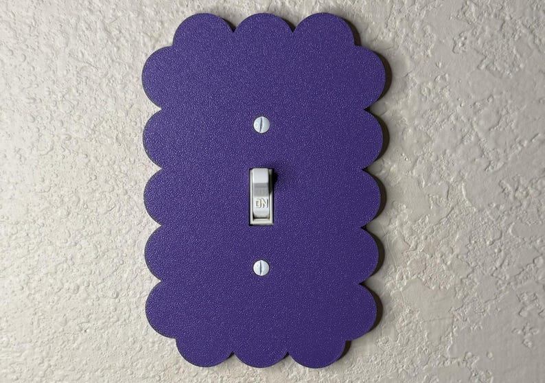 May include: A decorative purple light switch cover with a scalloped edge. The cover has a standard toggle switch in the center, with two screws at the top and bottom. The switch is in the "ON" position.