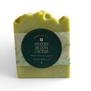 May include: A rectangular bar of light green artisan soap with a textured top. A dark green circular label wraps around the center, featuring the text: Emerald and Pearl, Desert Bloom Cactus, Fresh Cactus & Aloe Bar, Artisan Soap | 4 oz.