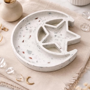 May include: A white terrazzo-style tray in a crescent moon shape with a star cutout. The tray has flecks of red, gray, and brown. The tray is on a beige linen cloth, with a small vase and candle in the background.