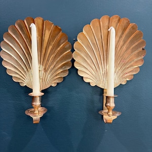 May include: A pair of matching wall-mounted candle sconces crafted from polished copper, each featuring a large, fan-shaped scallop shell backplate and a central cylindrical candle holder, with a single white taper candle inserted in each.