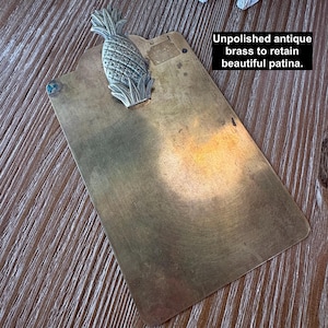 May include: Antique brass clipboard with a pineapple accent. The clipboard has a rectangular shape with rounded corners and a decorative pineapple design at the top. The text on the image reads: "Unpolished antique brass to retain beautiful patina."