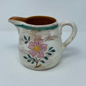 May include: A white ceramic creamer with a brown rim and a floral design. The flower is pink with a yellow center and green leaves.