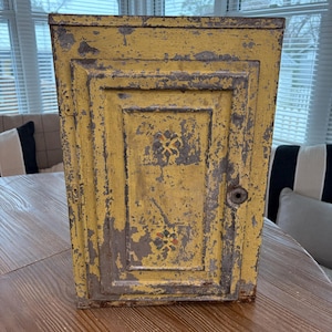 May include: A vintage yellow cabinet with a distressed finish. The cabinet has a rectangular shape with a single door and a keyhole. The paint is peeling, revealing gray underneath. Decorative floral patterns are visible on the door.