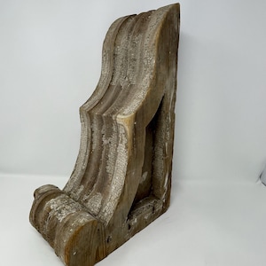 May include: A weathered wooden corbel with a curved design. The corbel has a natural wood finish and shows signs of age and wear.