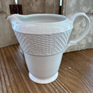 May include: A white ceramic creamer with a basket weave design around the middle. The creamer has a handle and a spout for pouring. The creamer sits on a small, round base. The creamer is a classic piece for serving cream or milk.