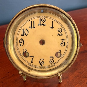 May include: Antique clock face with a cream-colored dial and black numerals. The clock face is housed in a gold-toned metal frame and has a small hole at the top. The text "Made by the New Haven Clock Co New Haven, Conn. U.S.A." is printed at the bottom.