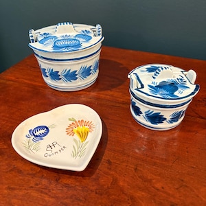 May include: Three-piece set of white ceramic kitchenware with blue floral designs. Includes two lidded containers and a heart-shaped dish with floral and text details. The set is displayed on a wooden surface.