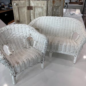 May include: A white wicker loveseat and armchair set. The furniture has a curved back and armrests with a woven design. The set is painted white and has a vintage aesthetic. The background includes antique doors.