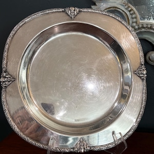 May include: A round silver-toned metal serving tray with a smooth, reflective center and a wide, decorative border. The rim features an intricate, embossed floral pattern with four distinct, ornate accents placed at equal intervals.