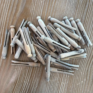 May include: A pile of wooden clothespins in various positions. The clothespins are made of light-colored wood and have a classic design with a spring-loaded mechanism. The clothespins are scattered on a light brown wooden surface.
