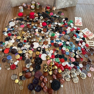 May include: A large collection of assorted sewing buttons in various shapes, sizes, and colors, including red, blue, black, white, gold, and brown. Several buttons are attached to two La Mode branded cardboard display cards with printed text.