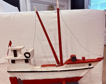 May include: A handcrafted wooden boat model, painted white with red accents. The boat features two red masts, a small red flag, and the text "MONT. NOTRE DAME." The model is displayed on a dark surface.
