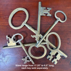 May include: A collection of antique brass keys of various sizes, arranged on a dark wooden surface. The keys have different head shapes, including oval and ornate designs. Sizes range from 17.25" to 8.5" long, each key sold separately.