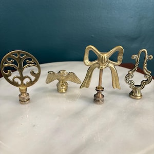 May include: A set of four brass lamp finials featuring distinct designs: a circular openwork scroll pattern, a small eagle with wings spread, a decorative bow shape on a thin rod, and an ornate vertical scroll piece with a textured base.