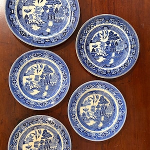 May include: A collection of five blue and white ceramic plates. Each plate features a detailed landscape scene with a pagoda, trees, and birds. The plates have a decorative border and are arranged on a wooden surface.