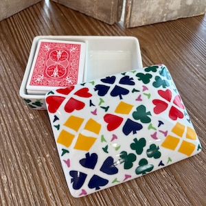 May include: A white ceramic playing card holder with a lid featuring a colorful pattern of hearts, spades, clubs, and diamonds. The holder contains a deck of red playing cards. The holder is on a wooden surface.