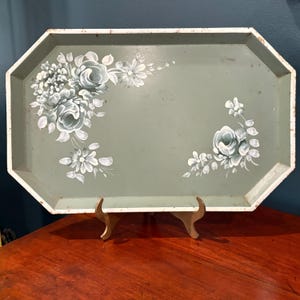 May include: An octagonal, vintage-style serving tray in a muted green hue, adorned with hand-painted white and gray floral designs. The tray has a white trim and is displayed on a wooden stand, showcasing its decorative appeal.