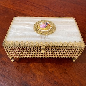 May include: Rectangular jewelry box with a creamy white lid and gold filigree detailing. The lid features a small, round, gold-framed porcelain cameo with a painted scene of two figures. The box has a gold clasp and rests on small feet.