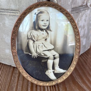 May include: Oval-shaped antique photo frame with a black and white portrait of a young girl. The frame is made of copper-colored metal with a decorative floral pattern around the edge. The girl is wearing a dress, socks, and shoes.