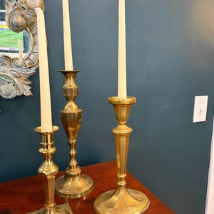May include: Three polished brass candlesticks of varying heights, each featuring a slender stem and a wide base. Each candlestick holds a single, tall, off-white taper candle. The brass surfaces exhibit a reflective, metallic gold-toned finish.