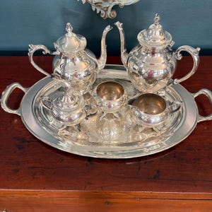 May include: A silver tea service set consisting of two teapots, a lidded sugar bowl, an open sugar bowl, and a creamer, all arranged on a large, oval silver serving tray with two handles. Each piece features a polished, reflective finish.