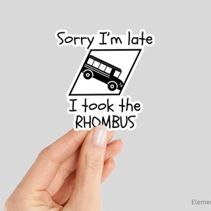 Peut inclure: Un autocollant noir et blanc avec le texte "Sorry I'm late I took the RHOMBUS." Le design présente un bus scolaire à l'intérieur d'un losange. L'autocollant est tenu par une main.