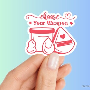 May include: Pink and white sticker with the text "choose your weapon" and a heart. The sticker features a makeup sponge, a makeup brush, and a jar of cream.