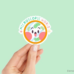 Peut inclure: Un autocollant rond avec une bordure orange et le texte "MICROSCOPIC WORLD" dans une police colorée. À l'intérieur du cercle se trouve une illustration de dessin animé d'une lame de microscope avec un visage souriant et des bactéries vertes et bleues.