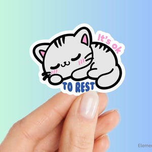 May include: A sticker featuring a sleeping cartoon cat with the text "It's ok to rest." The cat is white with black outlines and pink accents. The text is in pink and blue. The sticker is held by a hand.