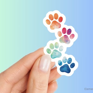 May include: Four colorful paw print stickers with a watercolor effect. The paw prints are arranged in a stack, with the largest paw print at the bottom and the smallest paw print at the top. The paw prints are in shades of orange, red, blue, and green.