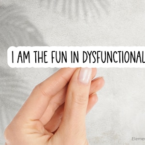 May include: A white sticker with the text "I AM THE FUN IN DYSFUNCTIONAL" in black, held by a hand. The sticker has a rounded rectangle shape. The background is a light gray.