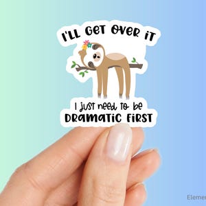 May include: A sticker featuring a cartoon sloth hanging from a branch, wearing a flower crown. The text reads "I'll get over it, I just need to be dramatic first." The sticker is held by a hand against a blue and green gradient background.