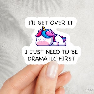 May include: A white unicorn with a pink and blue mane and horn is lying down with its eyes closed. The text "I'll get over it I just need to be dramatic first" is written on the sticker.