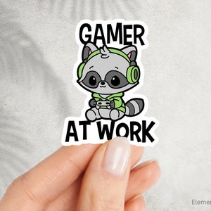 May include: A sticker depicting a cartoon raccoon in a green hoodie and headphones, holding a game controller. The words "GAMER AT WORK" are above and below the raccoon. The sticker is held by a hand.