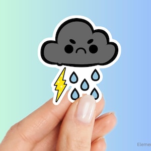 May include: A sticker featuring a cartoon cloud with an angry face, rain drops, and a yellow lightning bolt. The cloud is gray with a black outline. The rain drops are blue. The sticker is held by a person.
