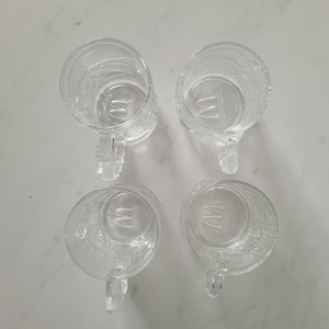 May include: Four clear glass mugs with handles, arranged in a square. Each mug has a textured exterior and a handle. The bottom of each mug features the McDonald's logo.