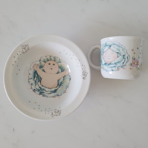 May include: A white ceramic bowl and mug with a Cabbage Patch Kid design. The bowl features a baby in a cabbage with a blue and green cabbage leaf design. The mug features a Cabbage Patch Kid face with a blue and green cabbage leaf design.