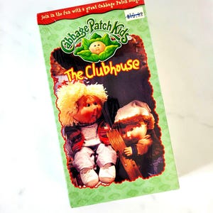 May include: A Cabbage Patch Kids VHS tape titled "The Clubhouse." The cover shows two dolls, one with blonde hair and the other with a winter hat, against a green background. The text "Join in the fun with 4 great Cabbage Patch Songs" is at the top.