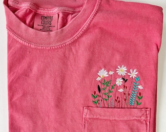 May include: A pink t-shirt with a pocket and embroidered white daisies and green leaves. The shirt has a tag that reads "Comfort Colors" and "Heavyweight Garment Dye 100% Cotton Made in Honduras".