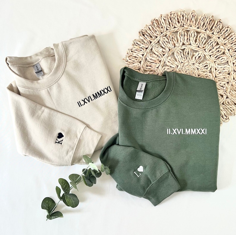 May include: Two sweatshirts, one beige and one green, with roman numeral dates and heart symbols embroidered on the sleeves. The beige sweatshirt has a black spade symbol on the sleeve and the green sweatshirt has a white heart symbol on the sleeve.