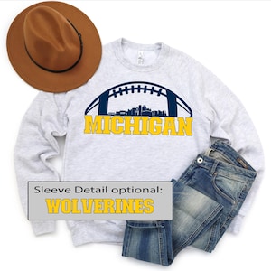 Puede incluir: Sudadera gris claro con un gráfico de fútbol americano y la palabra "MICHIGAN" en letras amarillas. El diseño incluye una silueta del horizonte de una ciudad. También hay unos vaqueros azules y un sombrero marrón en la imagen.