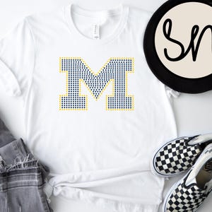 Peut inclure: T-shirt blanc avec un grand "M" en pointillés bleu marine et or. L'image comprend des lunettes de soleil bleues, un jean gris et des chaussures à enfiler à carreaux noirs et blancs. Un chapeau noir avec un cercle blanc et le logo "SN" est également présent.